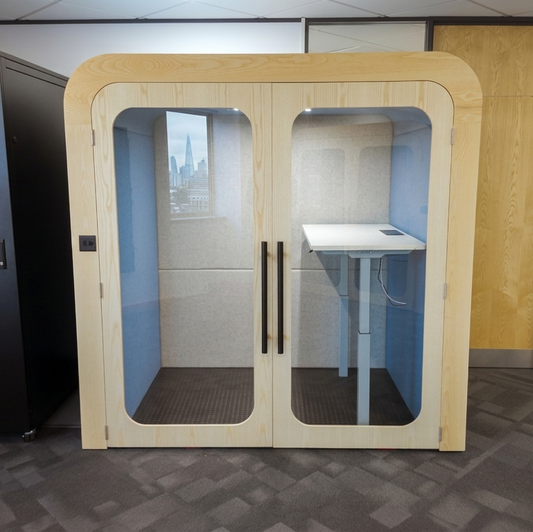 Small office pod with glass panels in a room with a fire extinguisher and bulletin board.